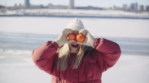 Young woman in white knitted hat and red winter jacket posing with two mandarins on the camera — Stock Video