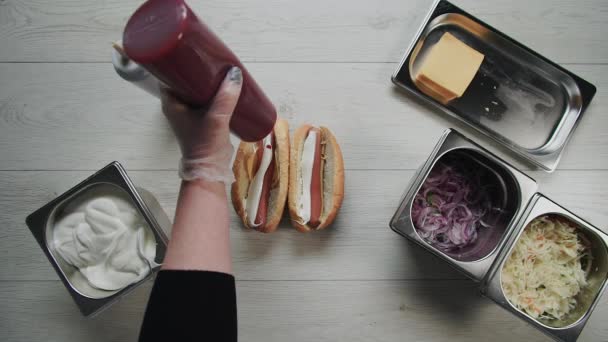 Bovenaanzicht van chef-kok handen in handschoenen kookt een hotdog, worst in het deeg. Chef zet souse in hotdog — Stockvideo