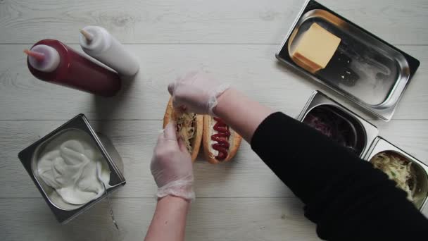 Vista dall'alto di chef mani in guanti cucina un hot dog, salsiccia nella pasta. Chef mette cavolo e cipolle sottaceto in hot dog — Video Stock