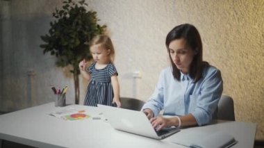 Çocuğu neredeyse sandalyeden düşerken genç anne dizüstü bilgisayarda çalışıyor.