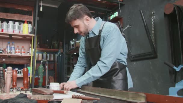 Der professionelle Gerber legt in der Werkstatt Kleister auf den Rand eines Bandes. Arbeitsprozess des Ledergürtels in der Lederwerkstatt. — Stockvideo