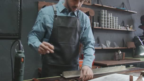Stilig man tillverkar ett brunt läderbälte med nål i läderverkstaden. Läderbältets arbetsprocess i läderverkstaden. — Stockvideo