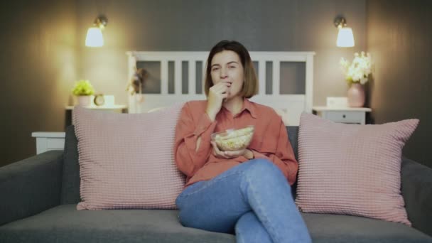Šťastná mladá žena sedí na gauči, jí popcorn, dívá se na televizi a směje se — Stock video