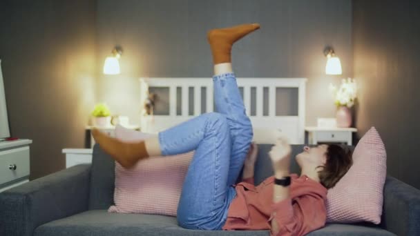 Mujer joven feliz acostada en el sofá, viendo películas de comedia y riendo — Vídeos de Stock