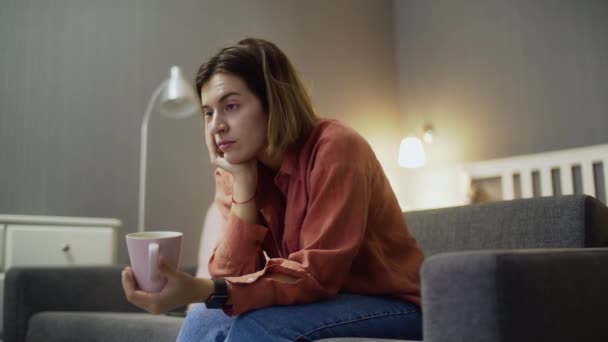 Joven mujer decepcionada con una taza de café sentada en el sofá y viendo la televisión — Vídeos de Stock