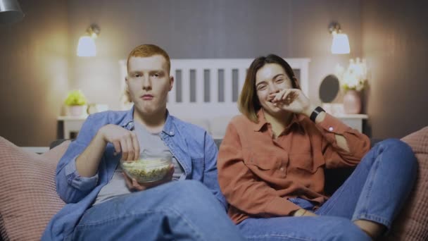 Щасливий молодий чоловік і жінка сидять на дивані, їдять попкорн, дивляться комедійний фільм і сміються — стокове відео
