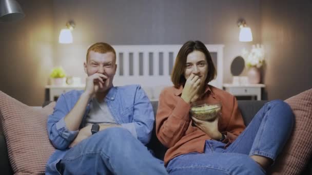 Boldog fiatal férfi és nő ülnek a kanapén, popcornt esznek, komédiás filmet néznek és nevetnek — Stock videók