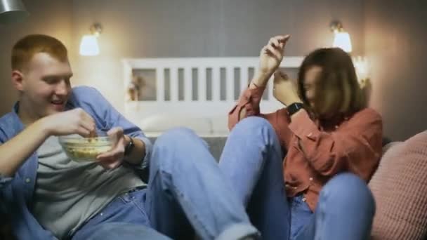 Feliz joven lanzando palomitas de maíz a la boca de las mujeres mientras se sienta en el sofá — Vídeos de Stock