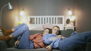 Uykulu genç adam ve kadın kanepede oturuyor ve televizyon seyrediyor.