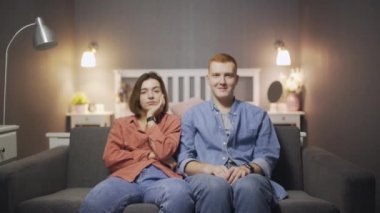 Mutlu genç adam kanepede oturup televizyon izlerken elini kadınların omzuna koyar.