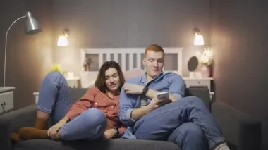 Genç bir kadın, erkek akıllı telefon kullanıp kanepede otururken televizyon seyrediyor.
