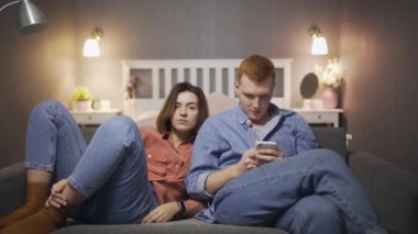 Genç bir kadın, erkek akıllı telefon kullanıp kanepede otururken televizyon seyrediyor.