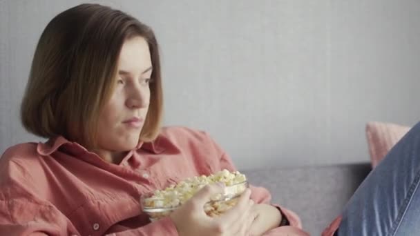 Молодая женщина сидит на диване, ест попкорн и смотрит телевизор — стоковое видео