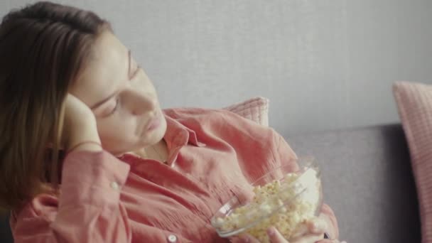Молодая женщина сидит на диване, ест попкорн, смотрит телевизор и падает снова — стоковое видео