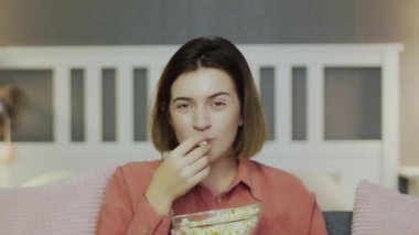 Kanepede oturan, patlamış mısır yiyen, televizyon seyreden ve gülen genç bir kadın.