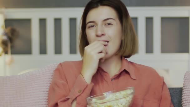 Glad ung kvinna sitter i soffan, äter popcorn, tittar på TV och skrattar — Stockvideo