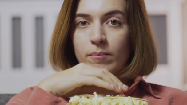 Närbild av ung kvinna sitter på soffan, äter popcorn och tittar på TV — Stockvideo