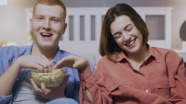 Joven chico feliz y chica viendo interesante película en la televisión junto con palomitas de maíz en la oscura sala de estar en el sofá. Cuarentena, pandemia, virus. Quédate en casa. — Vídeos de Stock