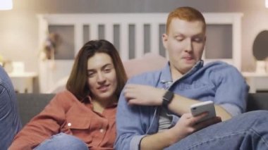 Genç bir kadın, erkek akıllı telefon kullanıp kanepede otururken televizyon seyrediyor.
