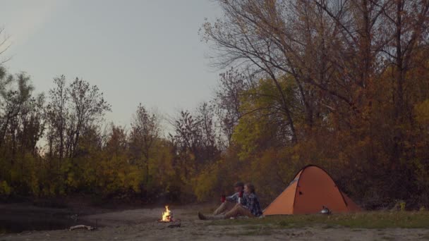 Couple of man and woman travelers sits near the bonfire and tent in an autumn forest — Stock Video