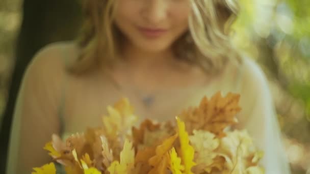 Красива усміхнена жінка тримає опале листя біля її обличчя і дивиться на камеру в осінньому парку на заході сонця — стокове відео