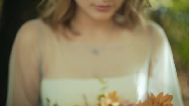 Bela mulher sorridente com olhos verdes segurando uma folhas caídas perto de seu rosto e olha para a câmera no parque de outono ao pôr do sol — Vídeo de Stock