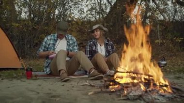 Bir çift erkek ve kadın gezgin şenlik ateşinin yanında oturur ve çubukların üzerinde şekerleme hazırlarlar.