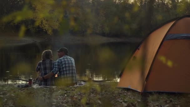 Back view of man and woman travelers sits hugging near the tent and river — Stock Video