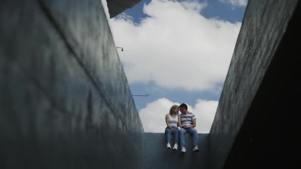 Joven pareja atractiva en vaqueros azules se sienta en la pared de hormigón — Vídeos de Stock