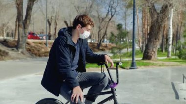 Tıbbi maskeli genç bir BMX sürücüsü bisiklette oturuyor ve kask takıyor.