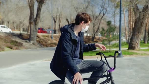 Un joven jinete de bmx con máscara médica sentado en una bicicleta y con un casco — Vídeos de Stock