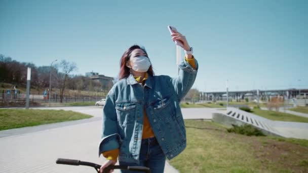 Ung asiatisk kvinna bloggare i en jeans jacka och medicinsk mask promenader med skotern och spela in video av dem själv i en sommarpark — Stockvideo