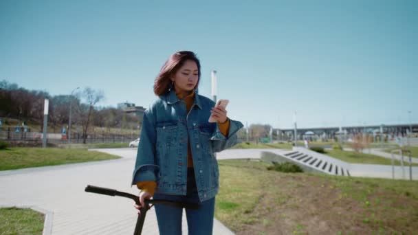 Jeune femme asiatique blogueur dans un jean veste promenades avec le scooter et enregistrer la vidéo d'eux-mêmes dans un parc d'été — Video