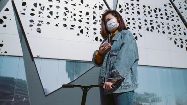 Молода азіатка в джинсовій куртці і медичній масці стоїть з скутером і записує голосове повідомлення в літньому парку — стокове відео
