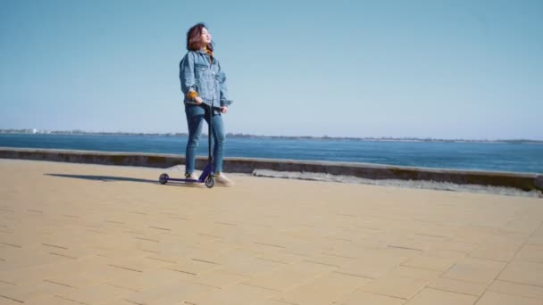 Jonge aziatische vrouw in een jeans jas loopt met de scooter in de buurt van de rivier — Stockvideo