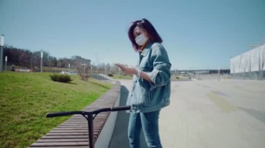 Tıbbi maskeli genç Asyalı kadın akıllı telefonu cebine koyar ve scooter 'a binmeye başlar.