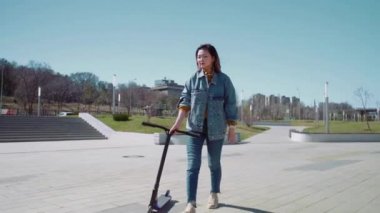 Genç Asyalı kadın yaz parkında scooterla yürüyor.
