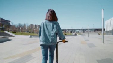 Kot ceketli genç bir kadının yaz parkında scooter sürüşünün arka görüntüsü.