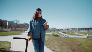 Tıbbi maskeli genç Asyalı kadın scooterıyla yürüyor ve yaz parkında akıllı telefon kullanıyor.