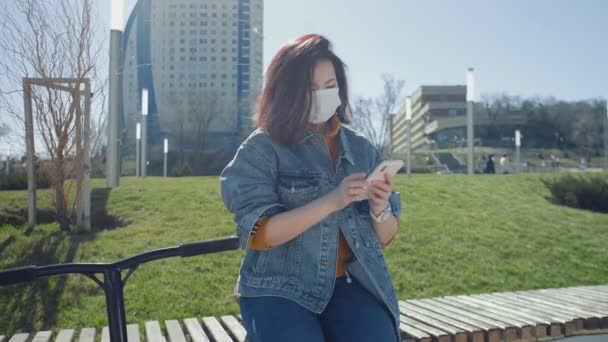 医療用マスクの若いアジアの女性がベンチに座って、夏の公園でスマートフォンを使用しています — ストック動画