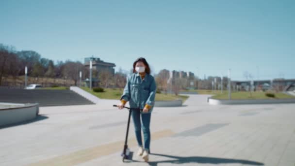 Junge Asiatin in medizinischer Maske spaziert mit Tretroller durch Sommerpark — Stockvideo