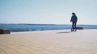 Kot ceketli genç Asyalı kadın nehir kıyısında scooter kullanıyor.