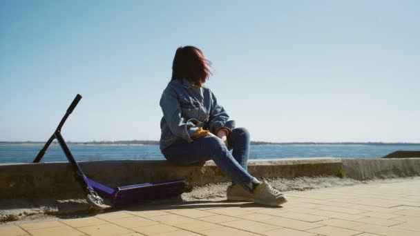 Молодая азиатка в джинсовой куртке сидит на границе со скутером у реки — стоковое видео