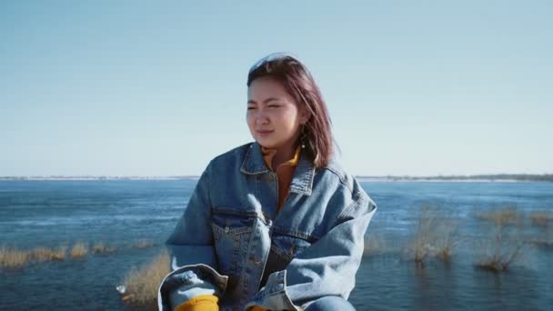 Jonge aziatische vrouw in een jeans jas hoesten terwijl zitten op de oever van de rivier — Stockvideo