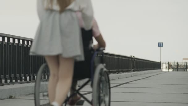 Инвалид в инвалидном кресле и его девушка, идущая по набережной — стоковое видео