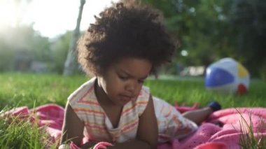 Afro-Amerikalı kızın yakın çekimleri. Parkta, yatak örtüsünün üzerinde uzanıyor ve telefonda çizgi film izliyor.