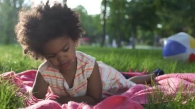 Afro-Amerikalı kızın yakın çekimleri. Parkta, yatak örtüsünün üzerinde uzanıyor ve telefonda çizgi film izliyor.