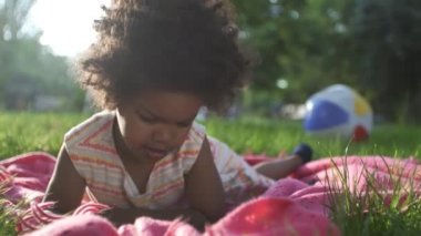 Afro-Amerikalı kızın yakın çekimleri. Parkta, yatak örtüsünün üzerinde uzanıyor ve telefonda çizgi film izliyor.