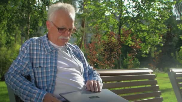 Літній чоловік сидить на лавці з ноутбуком в саду . — стокове відео