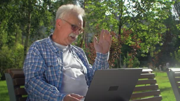 Naštvaný starší muž mluví skypem, sedí na lavičce v zahradě. Důchodce má na klíně laptop. Mává rukou, někomu nadává — Stock video